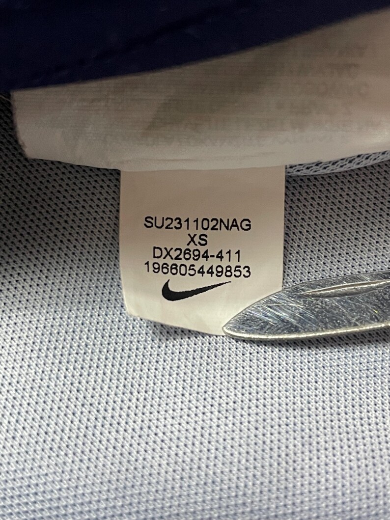 May include: A white clothing label sewn onto a light blue textured fabric. The label displays the text SU231102NAG, XS, DX2694-411, 196605449853, and a black Nike swoosh logo. A metallic object is partially visible at the bottom right of the label.