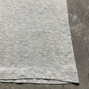 Puede incluir: Primer plano de una camiseta gris jaspeada. La tela tiene un aspecto texturizado con sutiles variaciones de color. Hay algunas peque&ntilde;as manchas cerca de la parte inferior de la camiseta. El dobladillo inferior est&aacute; doblado.