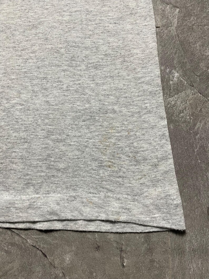 Puede incluir: Primer plano de una camiseta gris jaspeada. La tela tiene un aspecto texturizado con sutiles variaciones de color. Hay algunas peque&ntilde;as manchas cerca de la parte inferior de la camiseta. El dobladillo inferior est&aacute; doblado.