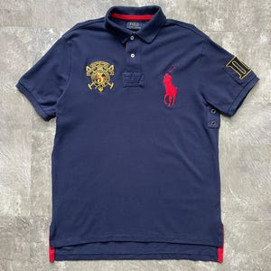 May include: A navy blue short-sleeve polo shirt featuring a red embroidered horse logo on the chest, a yellow and black crest patch with the number 3, and a large number 3 patch on the sleeve. The shirt has a collar with red trim and two buttons.