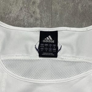 Puede incluir: Camiseta sin mangas deportiva blanca con el logo de Adidas en azul marino. La etiqueta muestra el logo de Adidas e informaci&oacute;n de tallas. La camiseta tiene un panel de malla y est&aacute; fabricada en China.