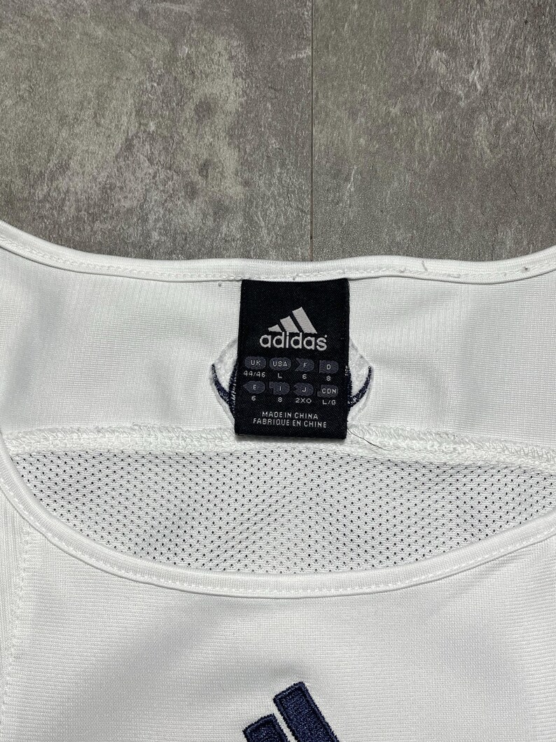 Puede incluir: Camiseta sin mangas deportiva blanca con el logo de Adidas en azul marino. La etiqueta muestra el logo de Adidas e informaci&oacute;n de tallas. La camiseta tiene un panel de malla y est&aacute; fabricada en China.