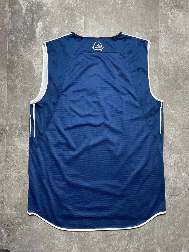K&ouml;nnte beinhalten: Ein &auml;rmelloses, marineblaues Sporttrikot mit wei&szlig;en Zierleisten und Akzenten. Das Trikot hat einen Mesh-R&uuml;ckeneinsatz und das Adidas-Logo auf dem oberen R&uuml;cken. Es hat einen gebogenen Saum und einen Rundhalsausschnitt.