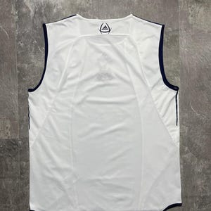 Puede incluir: Camiseta deportiva blanca sin mangas con ribete azul marino. La parte trasera de la camiseta presenta un logotipo azul marino en el cuello y un dise&ntilde;o de panel de malla. La camiseta est&aacute; hecha de una tela ligera y transpirable.