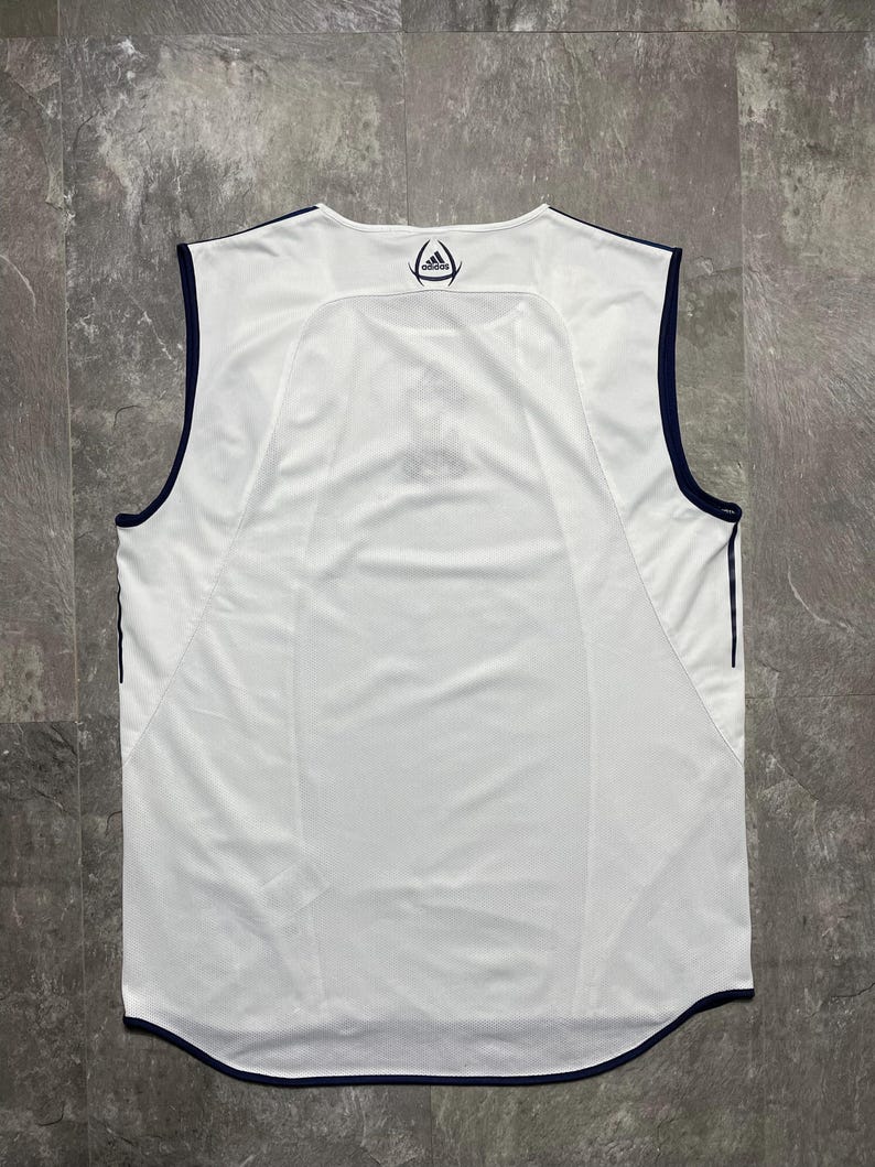 Puede incluir: Camiseta deportiva blanca sin mangas con ribete azul marino. La parte trasera de la camiseta presenta un logotipo azul marino en el cuello y un dise&ntilde;o de panel de malla. La camiseta est&aacute; hecha de una tela ligera y transpirable.