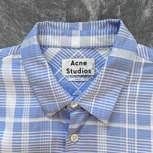 May include: A light blue and white plaid button-down shirt with a pointed collar. The interior neck label reads Acne Studios with the text Size 48 and Slim Fit printed below. The shirt features a front button placket with two visible white buttons.