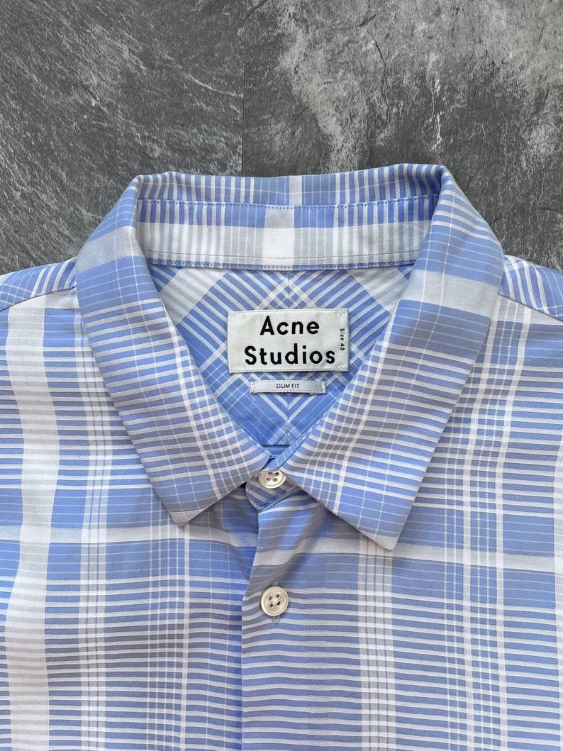 May include: A light blue and white plaid button-down shirt with a pointed collar. The interior neck label reads Acne Studios with the text Size 48 and Slim Fit printed below. The shirt features a front button placket with two visible white buttons.