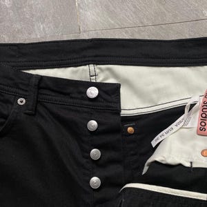 May include: Close-up of black denim jeans with silver button closure. The jeans have a white interior and a tag that reads "Acne Studios". The jeans are size 34/30.