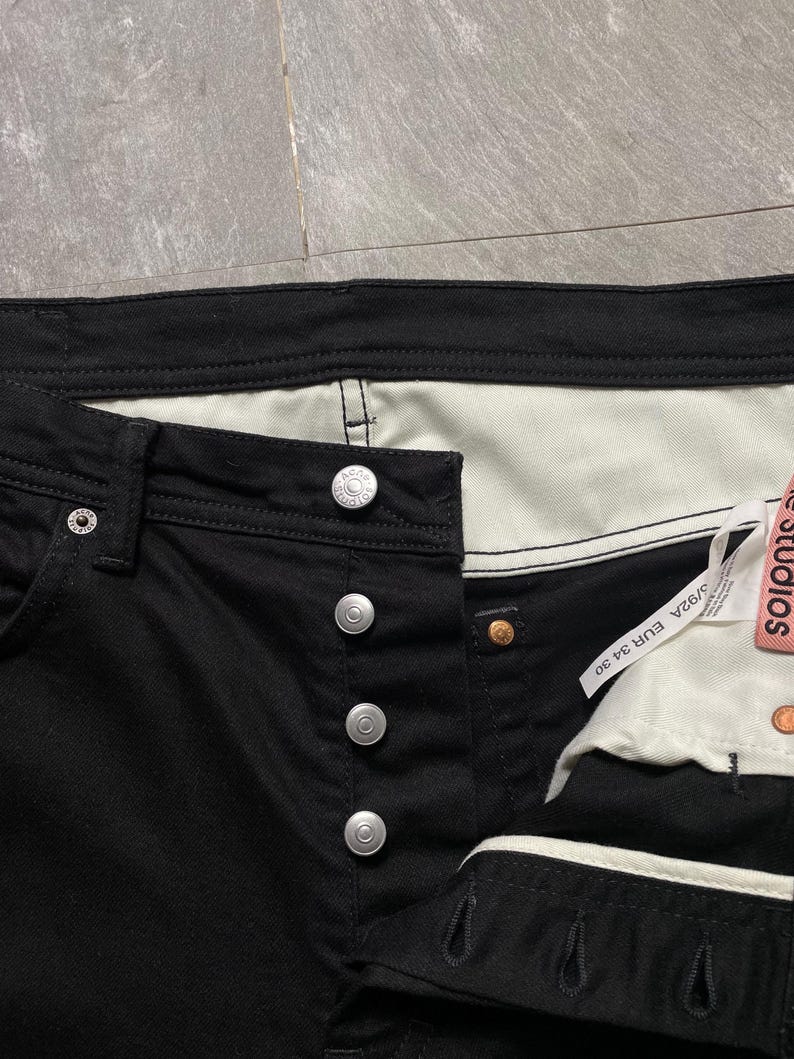 May include: Close-up of black denim jeans with silver button closure. The jeans have a white interior and a tag that reads "Acne Studios". The jeans are size 34/30.
