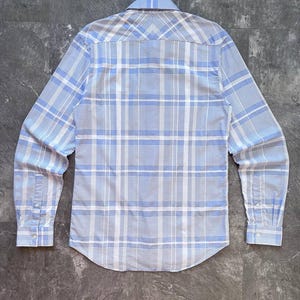 May include: A long-sleeve button-down shirt featuring a light blue and white plaid pattern. The shirt has a pointed collar, a back yoke with a chevron-style pattern alignment, and cuffs with two buttons each. The hem is curved at the bottom.