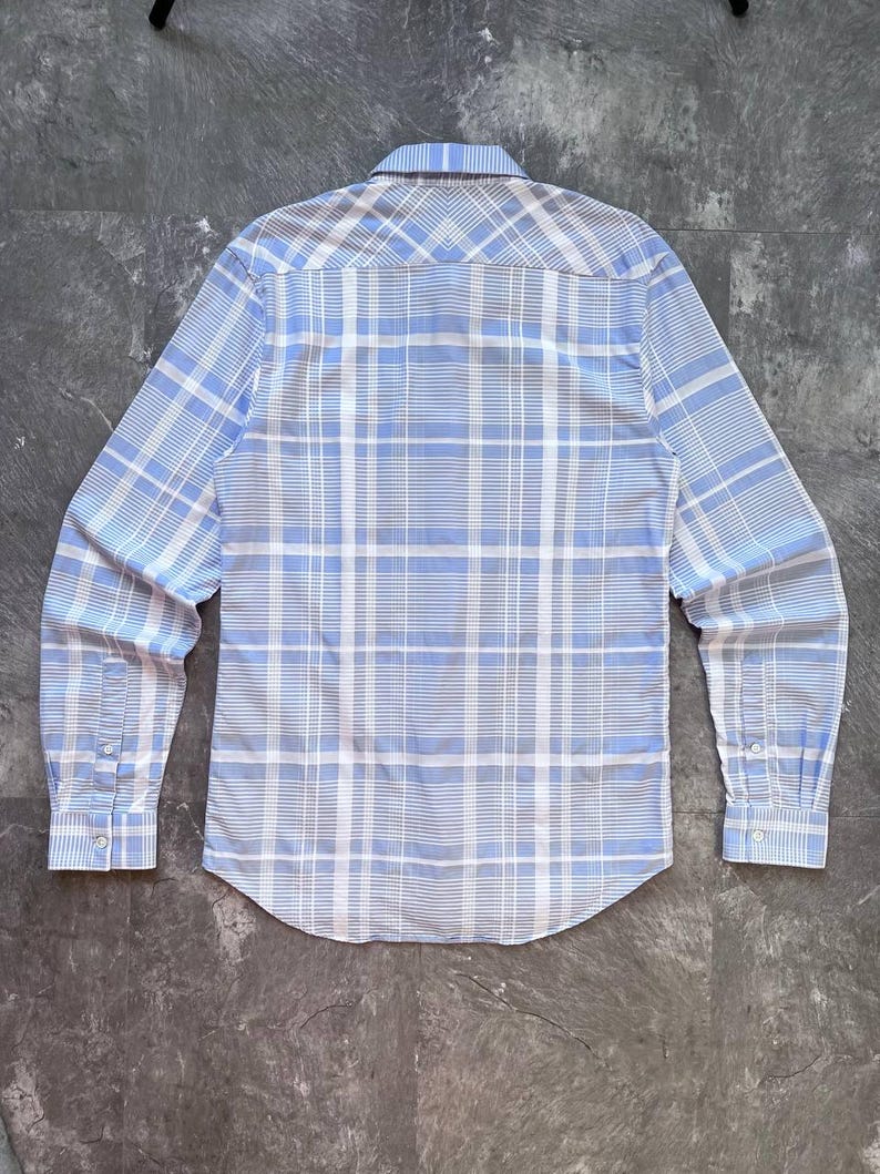 May include: A long-sleeve button-down shirt featuring a light blue and white plaid pattern. The shirt has a pointed collar, a back yoke with a chevron-style pattern alignment, and cuffs with two buttons each. The hem is curved at the bottom.