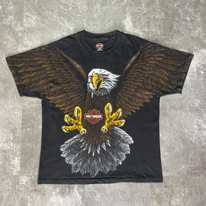 Puede incluir: Camiseta negra con un gran diseño de águila. El águila tiene la cabeza blanca, alas marrones y garras amarillas. El logotipo de Harley-Davidson es visible en el pecho. La camiseta es vintage y tiene un aspecto desgastado.
