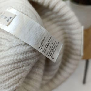 Może przedstawiać: Kremowa czapka z dzianiny o prążkowanym splocie z wewnętrzną metką. Na metce widnieje napis 56cm, FA-UX-HATS000024, C40044, Made in China oraz Shell 100% wool. Tekstura dzianiny jest wyraźnie widoczna na całej powierzchni czapki.