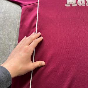 May include: A maroon athletic shirt with a white stripe down the side. The shirt features the words "Dr. Martens" in white, raised lettering. The fabric has a textured appearance.