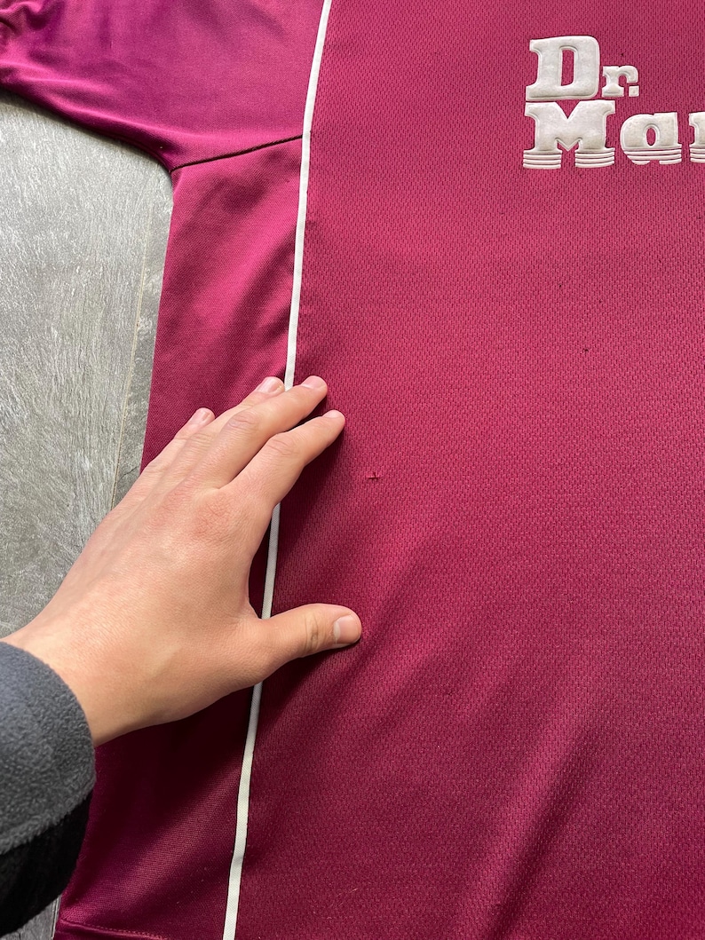 May include: A maroon athletic shirt with a white stripe down the side. The shirt features the words "Dr. Martens" in white, raised lettering. The fabric has a textured appearance.