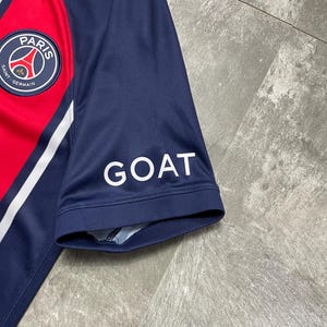 May include: A navy blue short-sleeved athletic jersey featuring a red vertical stripe, white trim, and the Paris Saint-Germain circular logo on the chest. The word GOAT is printed in white capital letters on the sleeve of the jersey.