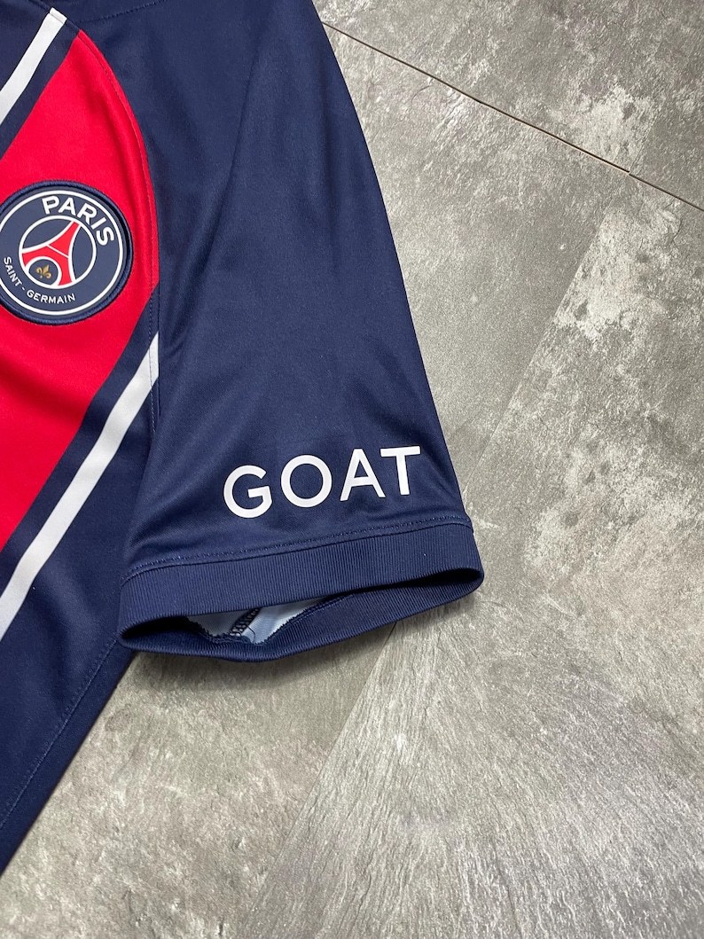 May include: A navy blue short-sleeved athletic jersey featuring a red vertical stripe, white trim, and the Paris Saint-Germain circular logo on the chest. The word GOAT is printed in white capital letters on the sleeve of the jersey.