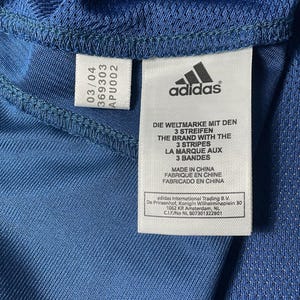 K&ouml;nnte beinhalten: Nahaufnahme des Etiketts eines blauen Sportshirts mit dem Adidas-Logo und Text in mehreren Sprachen. Das Etikett enth&auml;lt den Text "MADE IN CHINA" und andere Herstellungsinformationen.