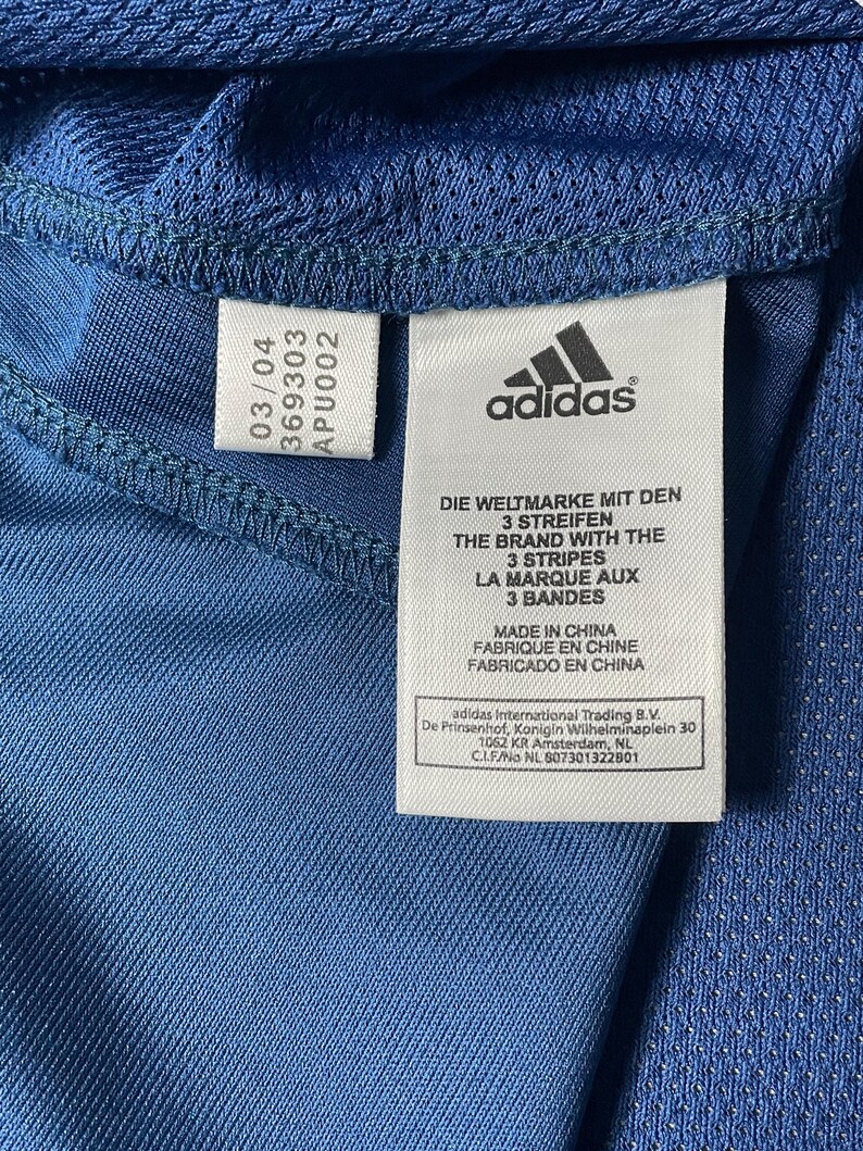 K&ouml;nnte beinhalten: Nahaufnahme des Etiketts eines blauen Sportshirts mit dem Adidas-Logo und Text in mehreren Sprachen. Das Etikett enth&auml;lt den Text "MADE IN CHINA" und andere Herstellungsinformationen.