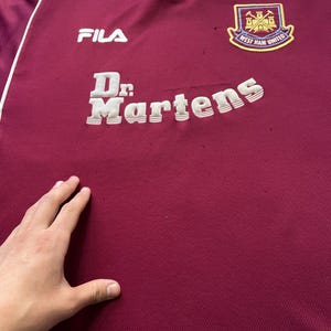 May include: Maroon and light blue vintage football shirt with the text "Dr. Martens" and the "West Ham United" logo. The shirt also features the "FILA" logo.