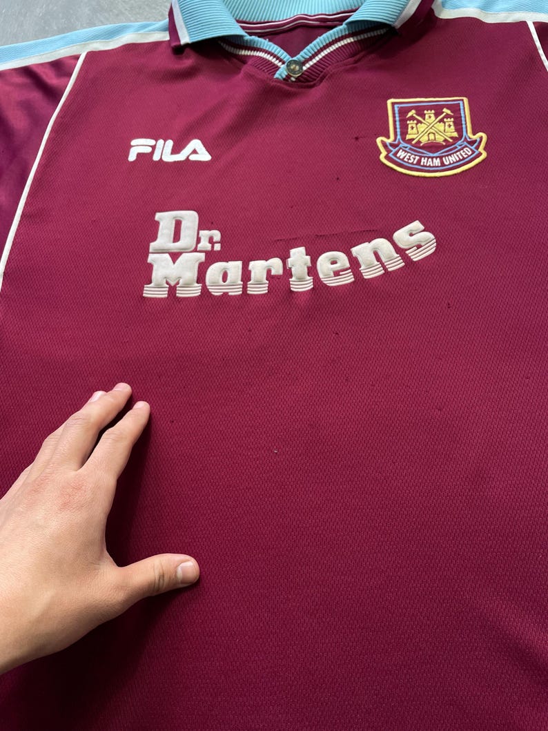 May include: Maroon and light blue vintage football shirt with the text "Dr. Martens" and the "West Ham United" logo. The shirt also features the "FILA" logo.