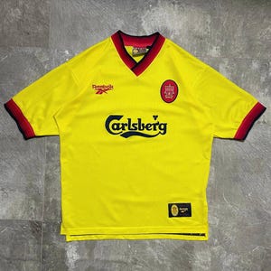 May include: A bright yellow short-sleeved jersey with a V-neck collar featuring red and black trim. The shirt displays the Reebok logo, a Liverpool Football Club crest, and the Carlsberg brand name in black text across the center chest area.