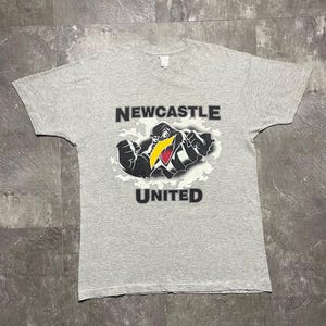Puede incluir: Camiseta gris jaspeado con la inscripci&oacute;n "NEWCASTLE UNITED" encima y debajo de una mascota de p&aacute;jaro de dibujos animados. El p&aacute;jaro es negro, blanco y amarillo, con los brazos flexionados. La camiseta es de cuello redondo.