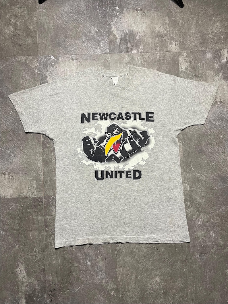 Puede incluir: Camiseta gris jaspeado con la inscripci&oacute;n "NEWCASTLE UNITED" encima y debajo de una mascota de p&aacute;jaro de dibujos animados. El p&aacute;jaro es negro, blanco y amarillo, con los brazos flexionados. La camiseta es de cuello redondo.