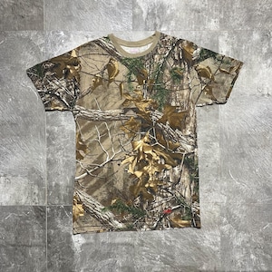 Op de afbeelding: Een camouflage T-shirt met korte mouwen met een realistisch bospatroon in bruin, groen en beige tinten. Het shirt heeft een ronde hals en een klein rood label bij de zoom. De stof lijkt licht van gewicht.