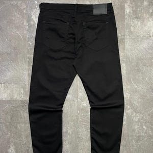 May include: Black denim jeans, seen from the rear. The trousers feature two back pockets and a leather patch on the waistband. The material looks to be of medium weight.