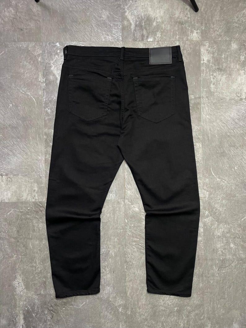 May include: Black denim jeans, seen from the rear. The trousers feature two back pockets and a leather patch on the waistband. The material looks to be of medium weight.