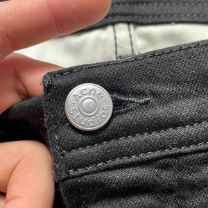 May include: Close-up of black denim fabric with a silver button. The button is embossed with the text "ACNE STUDIOS" in a circular design. The fabric shows detailed stitching.