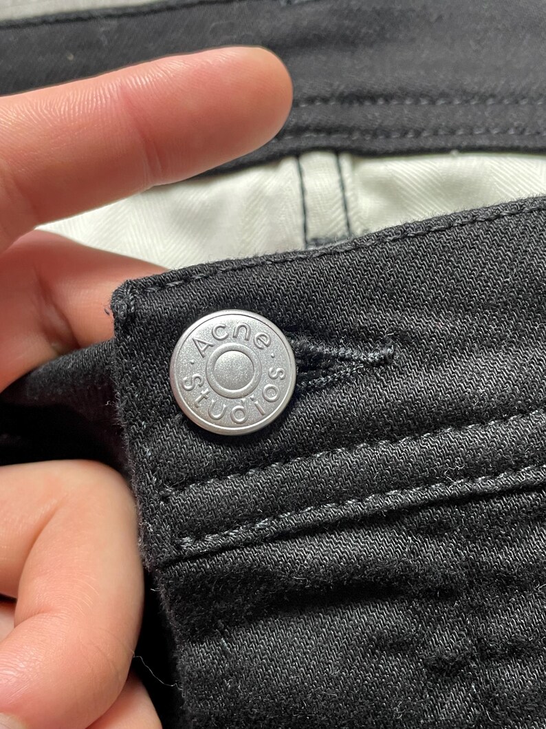 May include: Close-up of black denim fabric with a silver button. The button is embossed with the text "ACNE STUDIOS" in a circular design. The fabric shows detailed stitching.
