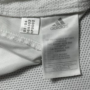 Puede incluir: Primer plano de una etiqueta de camiseta deportiva blanca con el logotipo de Adidas y texto. La etiqueta incluye el texto "MADE IN CHINA" y otros detalles. La camisa tiene una tela texturizada y transpirable.