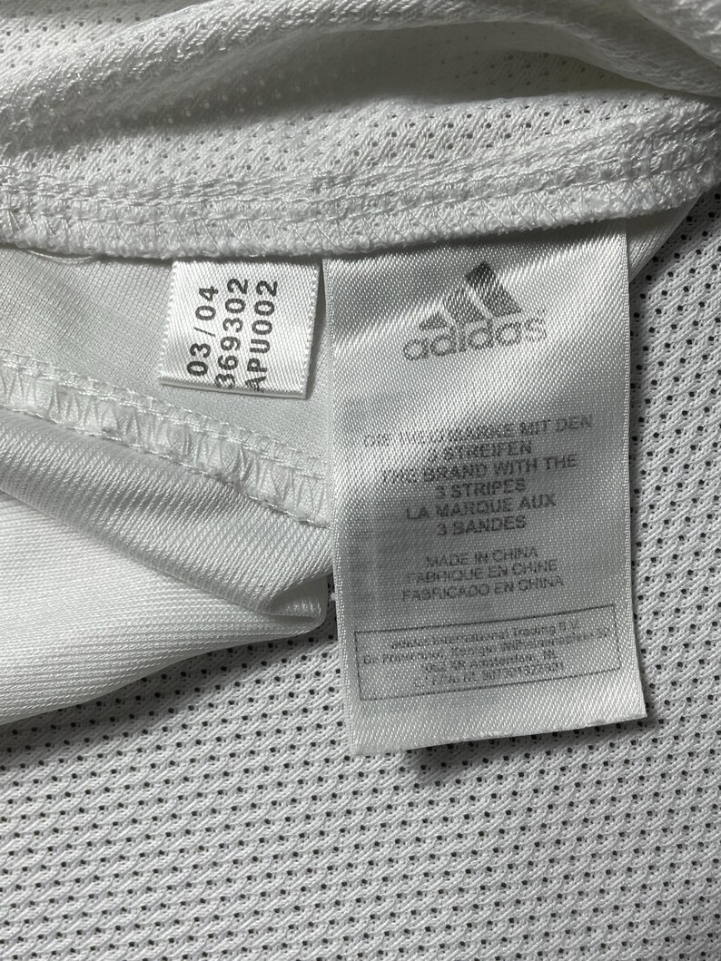 Puede incluir: Primer plano de una etiqueta de camiseta deportiva blanca con el logotipo de Adidas y texto. La etiqueta incluye el texto "MADE IN CHINA" y otros detalles. La camisa tiene una tela texturizada y transpirable.