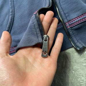 May include: Close-up of a blue zip-up hoodie with a silver zipper. The zipper pull has a horseshoe design. The hoodie has red stitching along the seams.