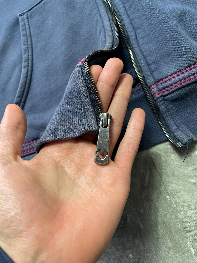 May include: Close-up of a blue zip-up hoodie with a silver zipper. The zipper pull has a horseshoe design. The hoodie has red stitching along the seams.