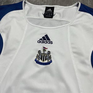 Puede incluir: Camiseta de f&uacute;tbol Adidas blanca y azul marino. La camiseta presenta el logo de Adidas y un escudo del equipo en el pecho. Tiene un panel de malla en el cuello y detalles en azul marino en los hombros.