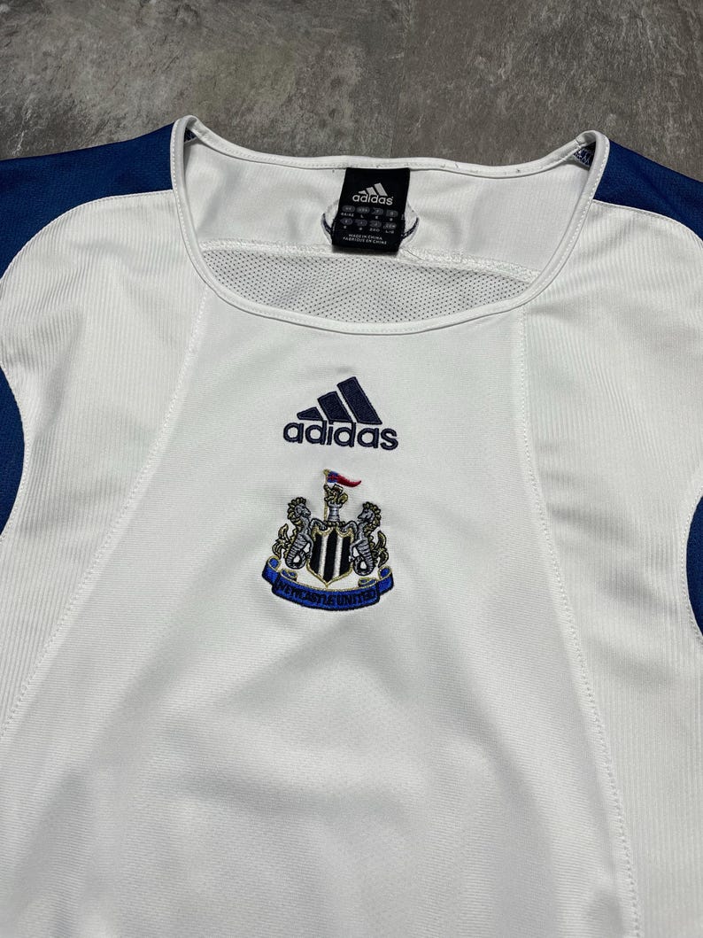Puede incluir: Camiseta de f&uacute;tbol Adidas blanca y azul marino. La camiseta presenta el logo de Adidas y un escudo del equipo en el pecho. Tiene un panel de malla en el cuello y detalles en azul marino en los hombros.