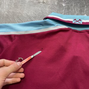 May include: Maroon polo shirt with light blue and white detailing on the collar and shoulders. The shirt features a textured weave and a small logo on the collar. A small pocket knife is held near the shirt.