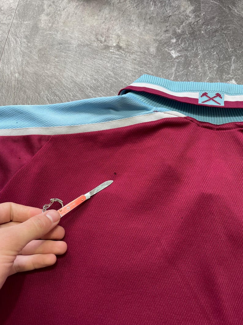 May include: Maroon polo shirt with light blue and white detailing on the collar and shoulders. The shirt features a textured weave and a small logo on the collar. A small pocket knife is held near the shirt.