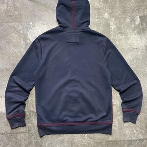 May include: Navy blue hoodie with a hood and long sleeves. The stitching is a contrasting red color. The hoodie has a small square patch on the back of the neck.