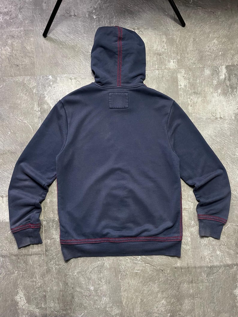 May include: Navy blue hoodie with a hood and long sleeves. The stitching is a contrasting red color. The hoodie has a small square patch on the back of the neck.