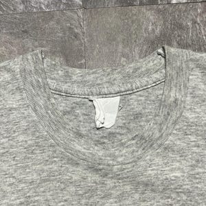 Puede incluir: Primer plano de una camiseta gris jaspeada. La camiseta tiene cuello redondo y una etiqueta visible. La tela tiene un aspecto texturizado.