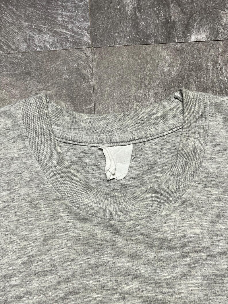 Puede incluir: Primer plano de una camiseta gris jaspeada. La camiseta tiene cuello redondo y una etiqueta visible. La tela tiene un aspecto texturizado.