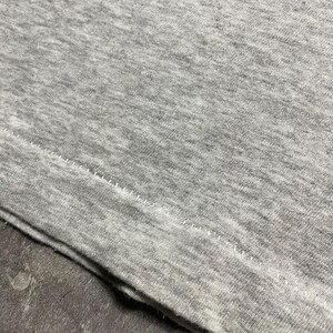 Puede incluir: Primer plano de una tela gris jaspeada, probablemente una camiseta, con una costura visible y puntadas a lo largo del borde. La tela tiene una textura suave y un aspecto ligeramente desgastado. La imagen se centra en los detalles del material.