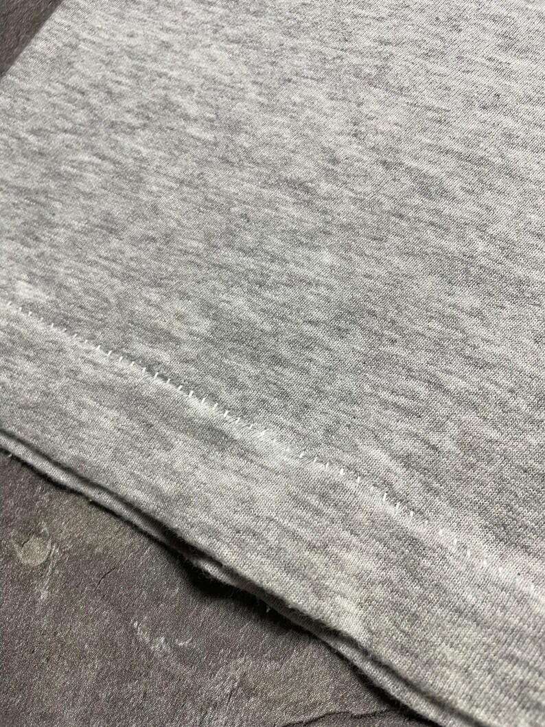 Puede incluir: Primer plano de una tela gris jaspeada, probablemente una camiseta, con una costura visible y puntadas a lo largo del borde. La tela tiene una textura suave y un aspecto ligeramente desgastado. La imagen se centra en los detalles del material.