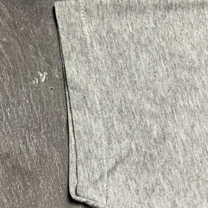 Puede incluir: Primer plano de una manga de camiseta gris. La tela tiene una textura jaspeada, con costuras visibles en el borde. La manga est&aacute; doblada en la parte inferior.