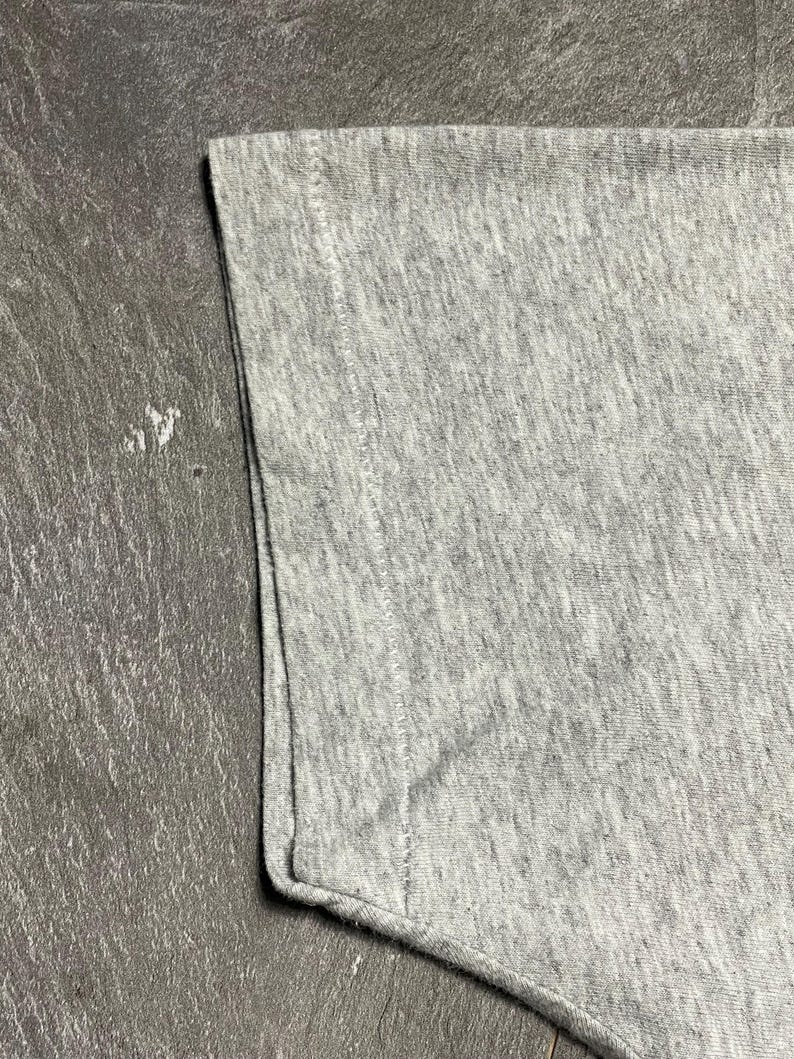 Puede incluir: Primer plano de una manga de camiseta gris. La tela tiene una textura jaspeada, con costuras visibles en el borde. La manga est&aacute; doblada en la parte inferior.
