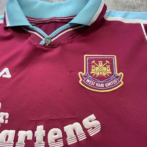 May include: Maroon and light blue West Ham United football shirt. Features a collared neckline, button closure, and the club crest. The Dr. Martens logo is also visible.